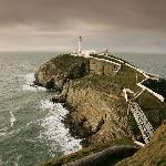 GeoMon Geopark - Wales. Site Name: South Stack on Holy Island, Anglesey. South Stack is only 10 minutes from the port of Holyhead and lies to the west of the sparkling white rocks of Anglesey's highest point, Holyhead Mountain. A Roman signal station sits on its highest point and an Iron-age village lies on its flanks. The magnificent green, schistose, folded rocks at the island and beneath the light-house are known world wide for their sheer beauty and the sandier competent beds of meta-sandstone are faulted. There are important sea-bird colonies on the cliffs and the maritime heath (heather and gorse) covers the striated quartzite on the mountain. This is probably the most picturesque and most-photographed site in Wales.