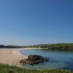 Geopark Shetland - Scotland. Site Name: St Ninian's Isle, Dunrossness. The tombolo connecting St Ninian's Isle to the mainland is one of the best examples of a sand tombolo in Europe. On a fine day it also provides one of the best amenity sites in Shetland. Tombolos are a feature of submerging coastlines. As the waves approach St Ninian's Isle from the west they bend around the island depositing the sediments they carry when they meet again on the east side of the island.